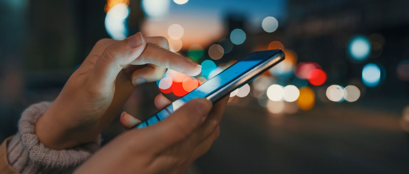 Hand with a smart phone in a dark. Female using her mobile phone, city skyline night light background. Girl pointing finger on screen smart phone on background illumination color light in night city